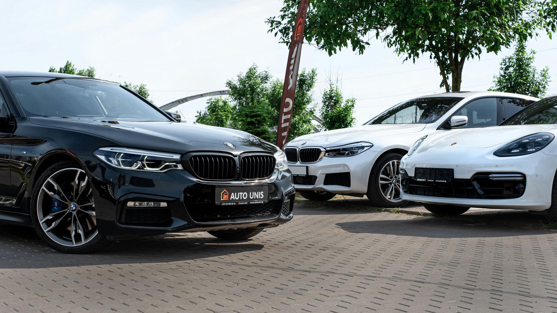 Black BMW, white BMW, and white Porsche parked outdoors on tiled pavement with trees in the background.