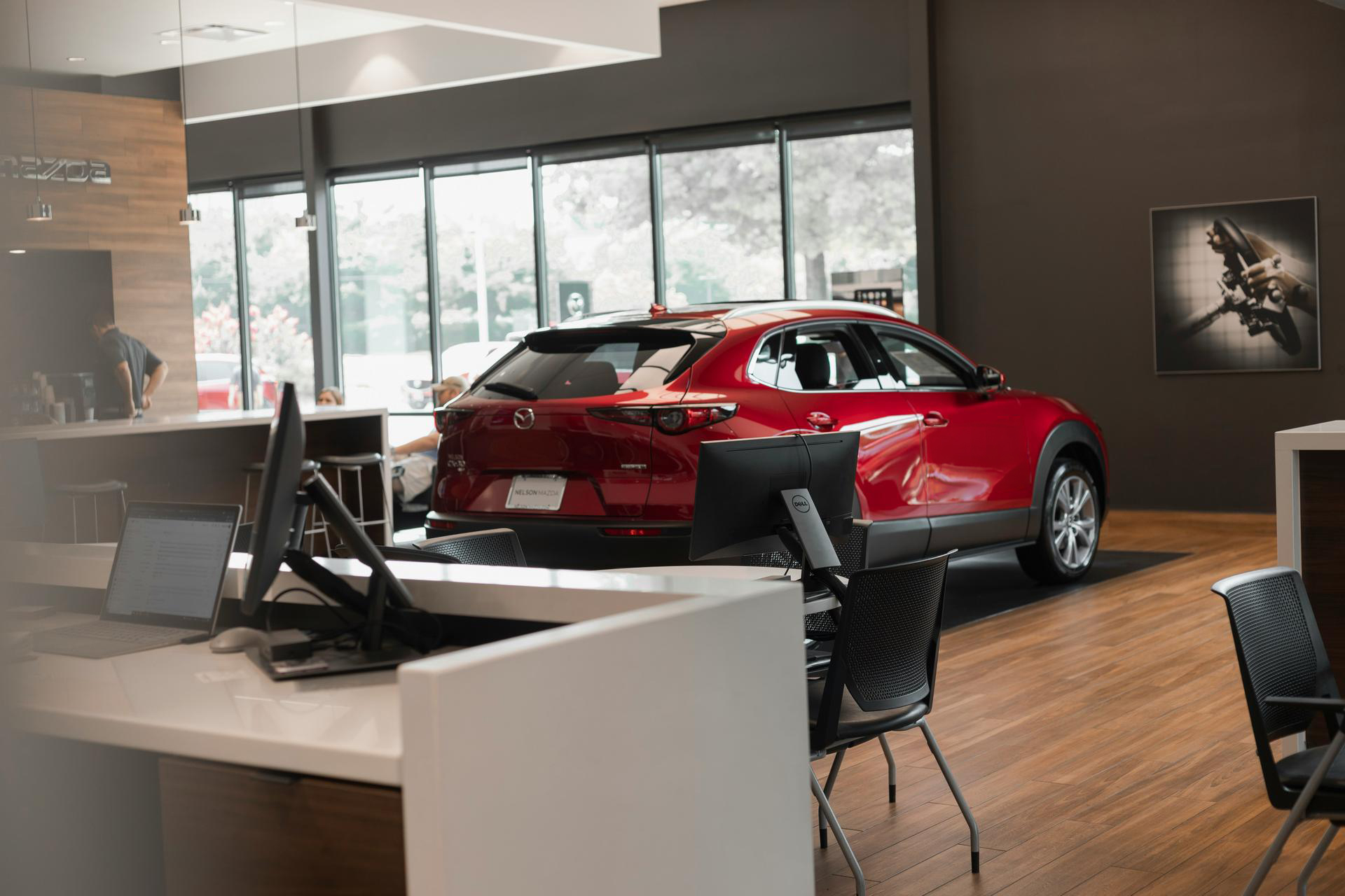 Red Mazda SUV displayed inside a modern car dealership with computer desks and chairs in the foreground.