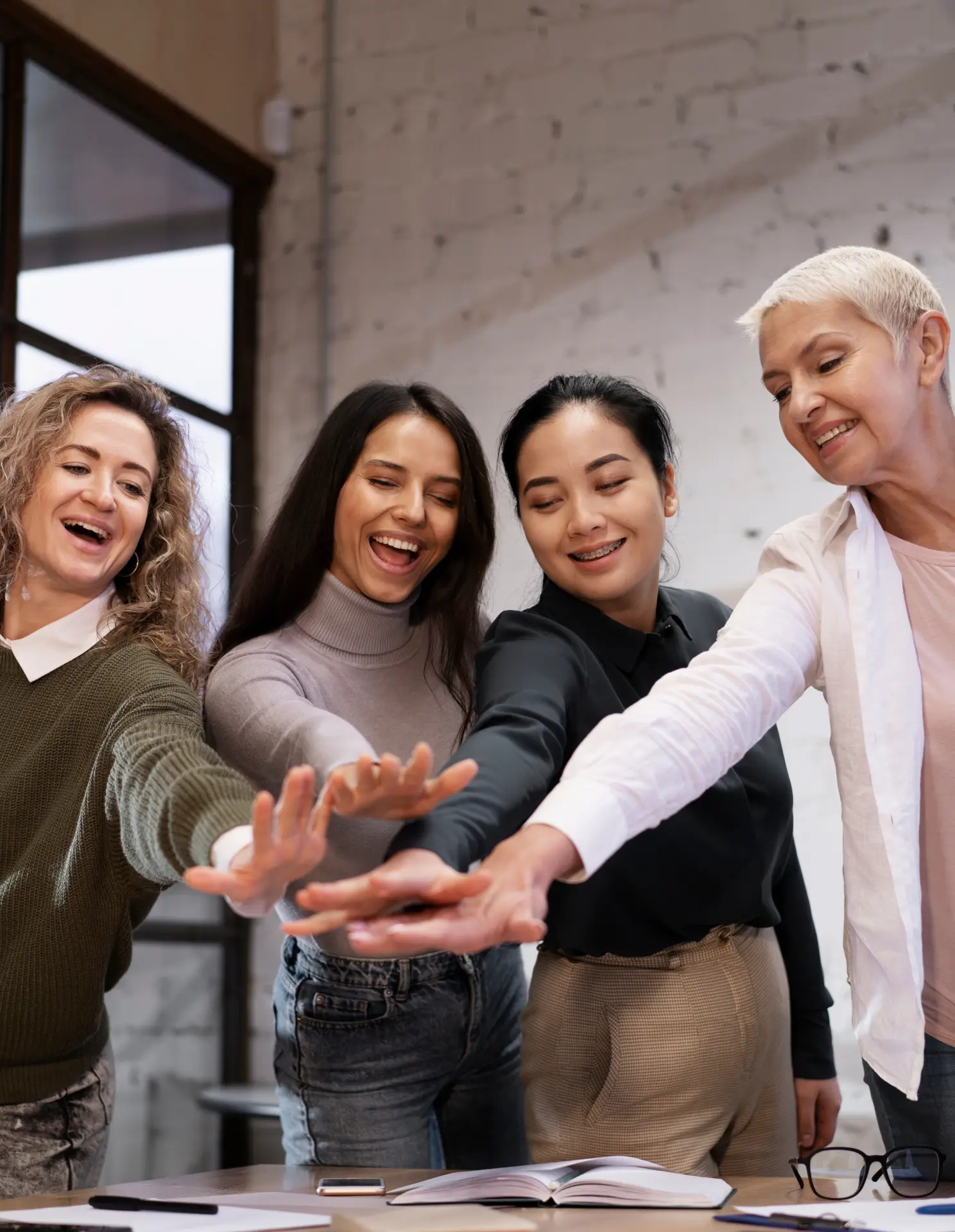 Four diverse women standing around a table with open books, smiling and stacking their hands together in a show of teamwork.