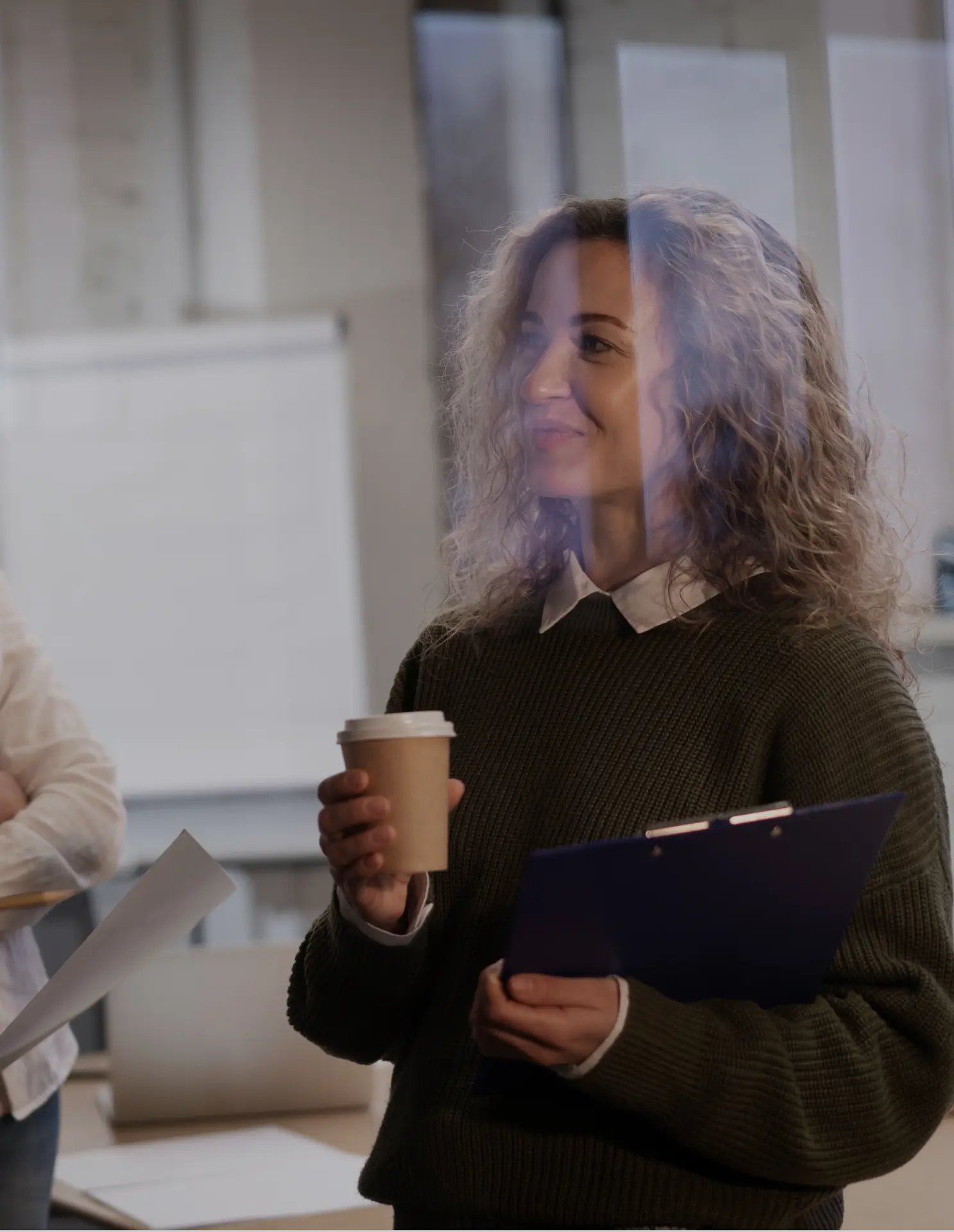 Young woman with curly hair holding a coffee cup and a clipboard, seen through a glass panel in an office setting.