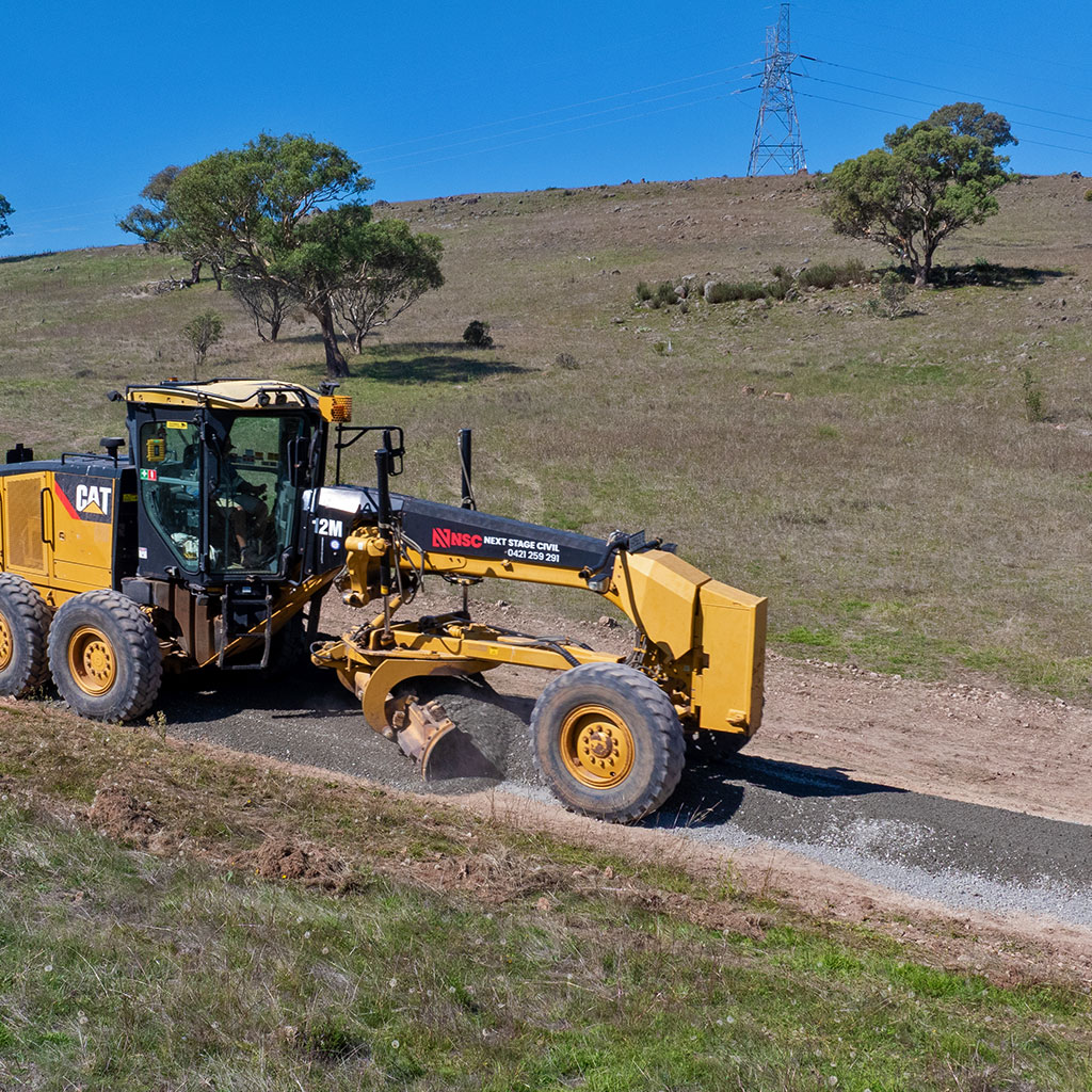 Yellow CAT motor grader leveling gravel on a rural dirt road under a clear blue sky.
