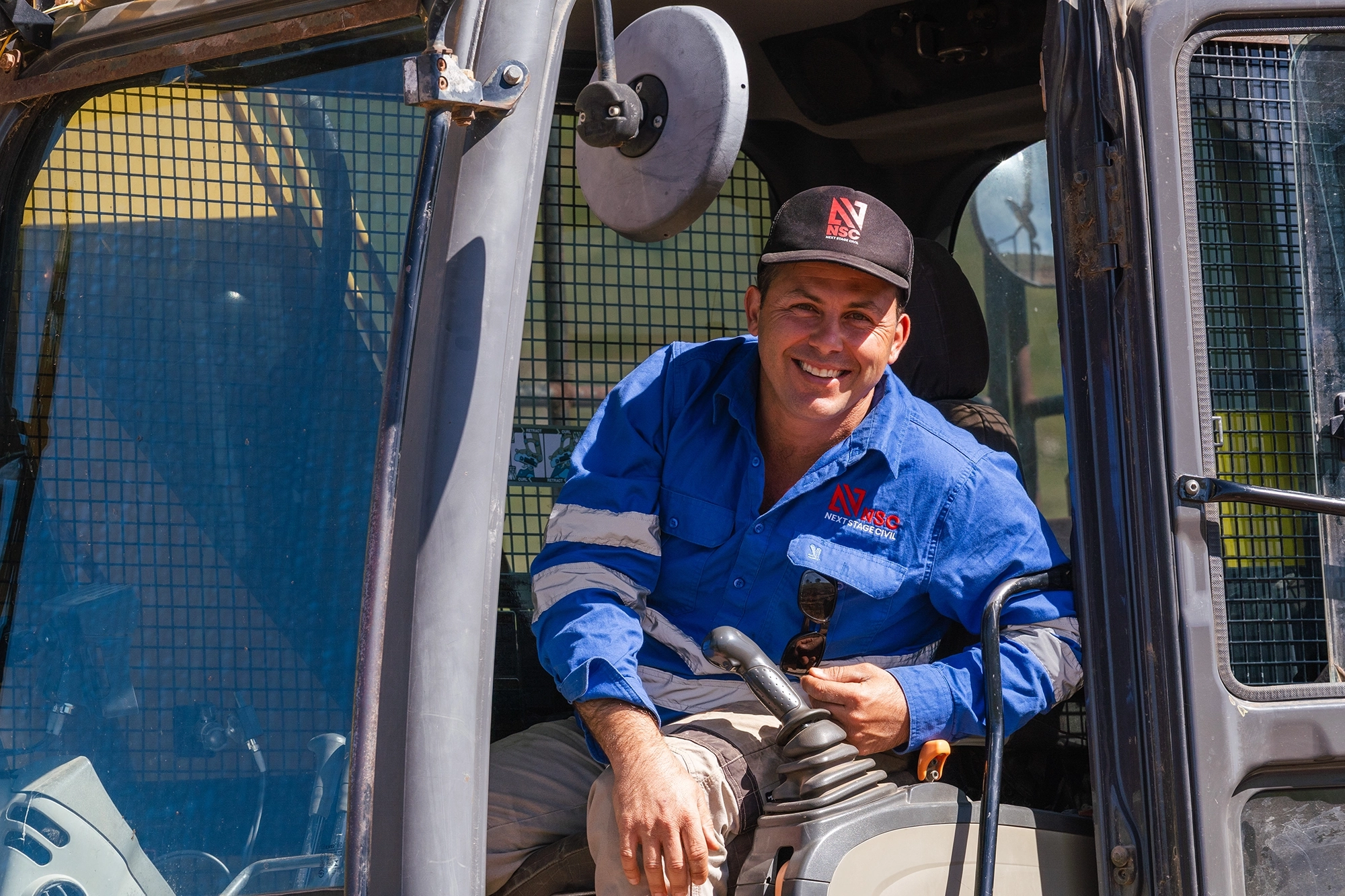 Daniel Grech, Director of Next Stage Civil, inside an excavator cab on site, wearing NSC branded uniform.