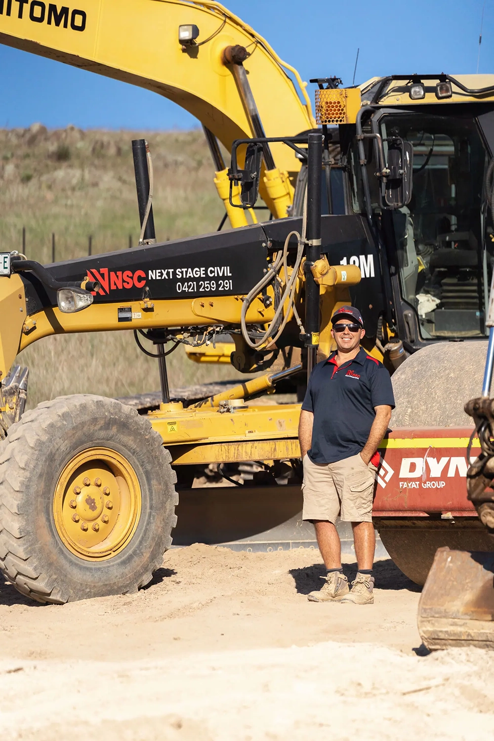 Man in sunglasses, navy and red polo, and beige shorts standing on a sandy construction site beside yellow heavy machinery with Next Stage Civil logo.