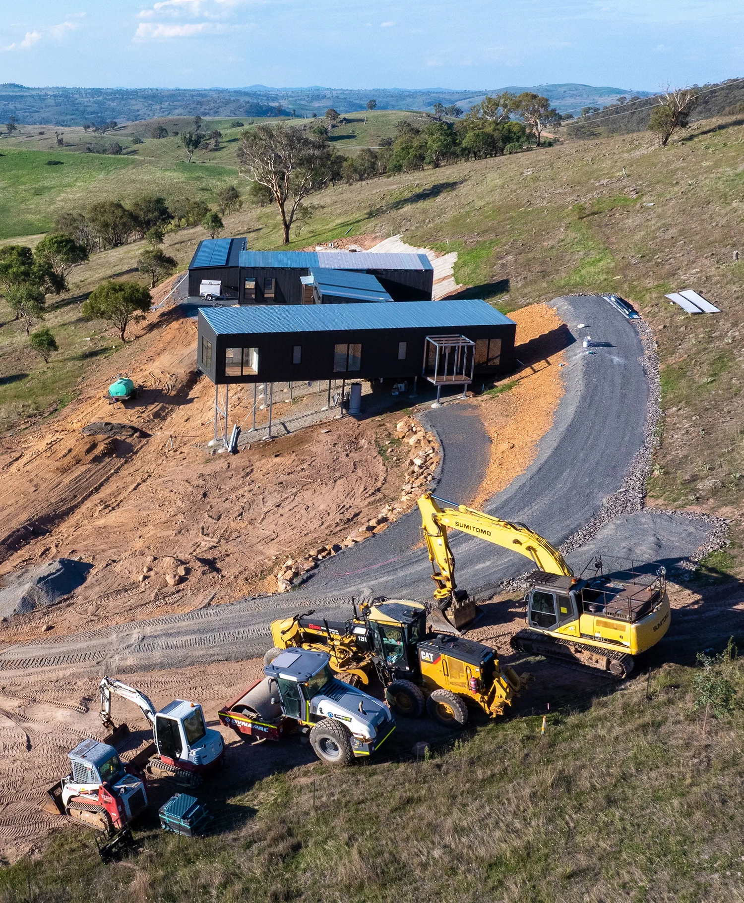 Aerial view of a modern black house on stilts on a hillside with construction vehicles and earthwork in progress around it.