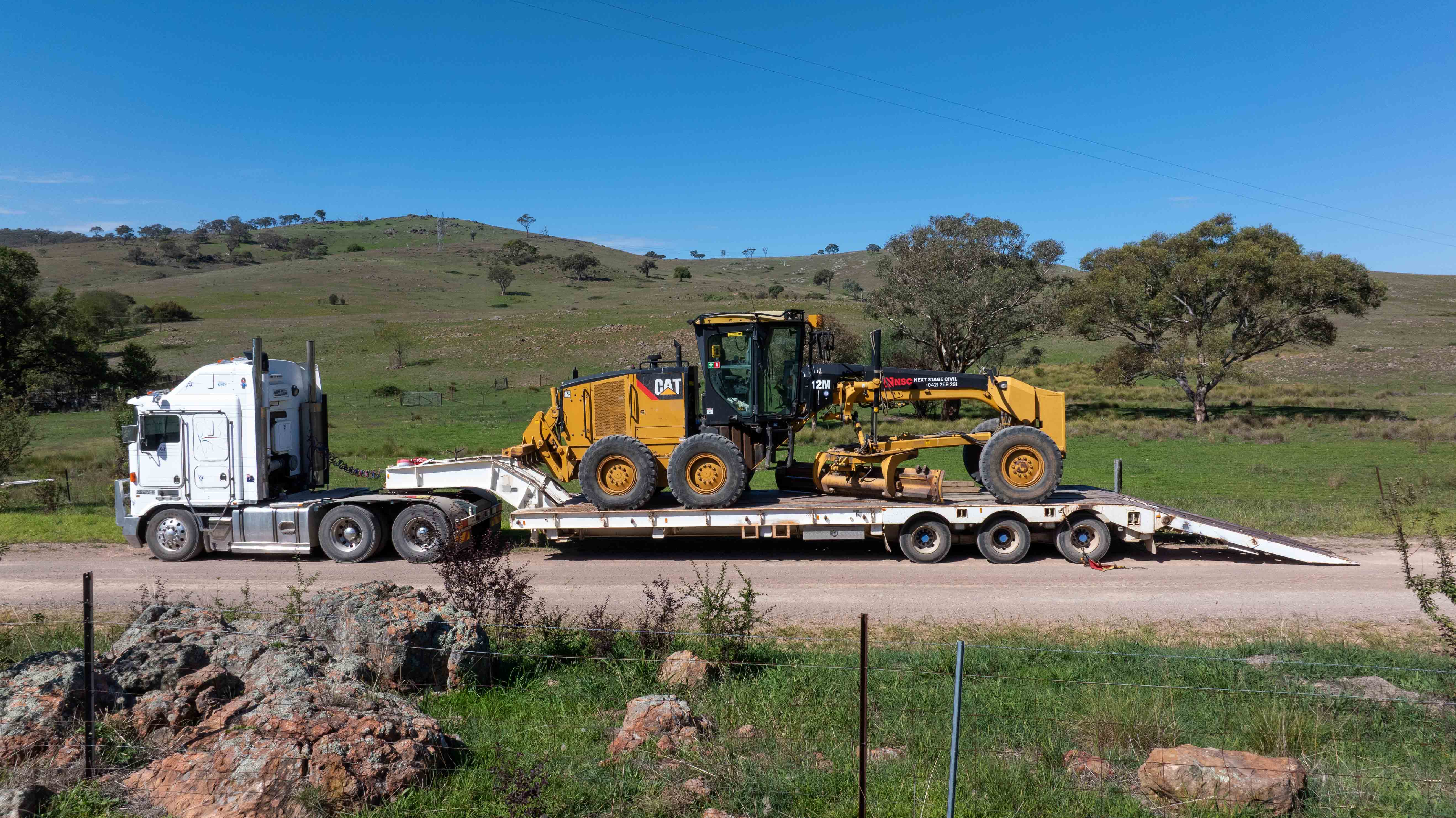 White semi-truck transporting yellow CAT 12M motor grader on a flatbed trailer on a rural road.