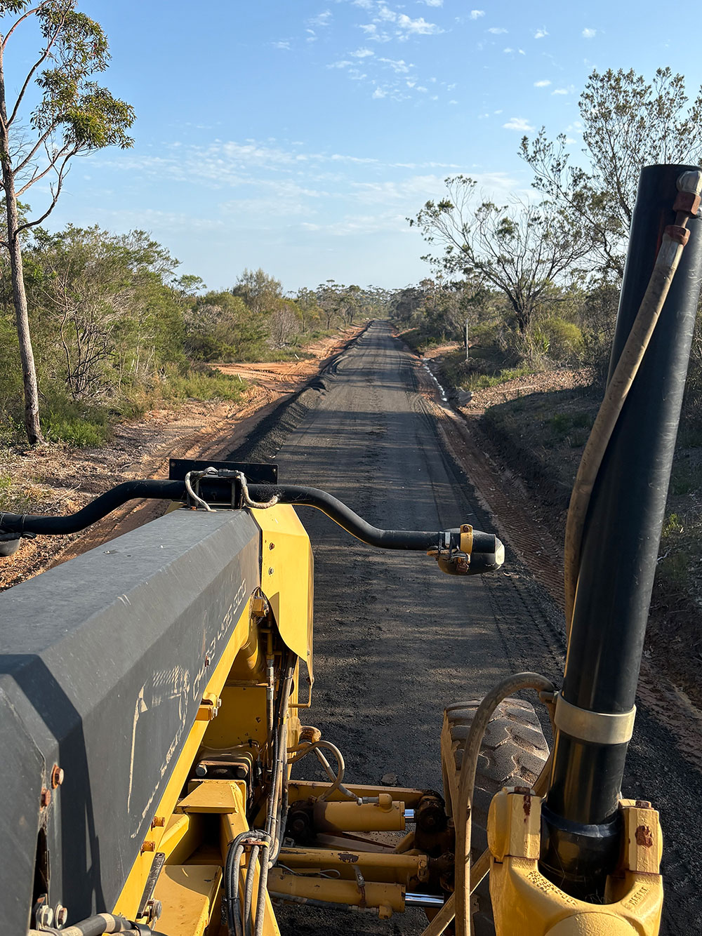 View from the cockpit of a yellow road grader working on a newly paved rural road surrounded by trees and shrubs under a blue sky.