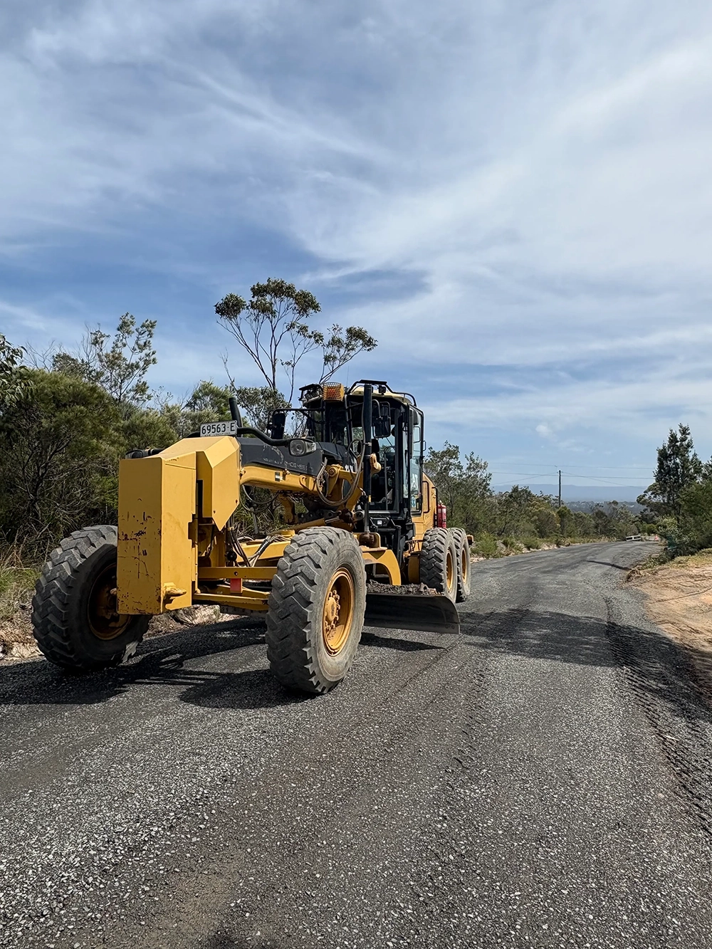 Yellow road grader parked on a gravel road under a partly cloudy sky with trees on the roadside.