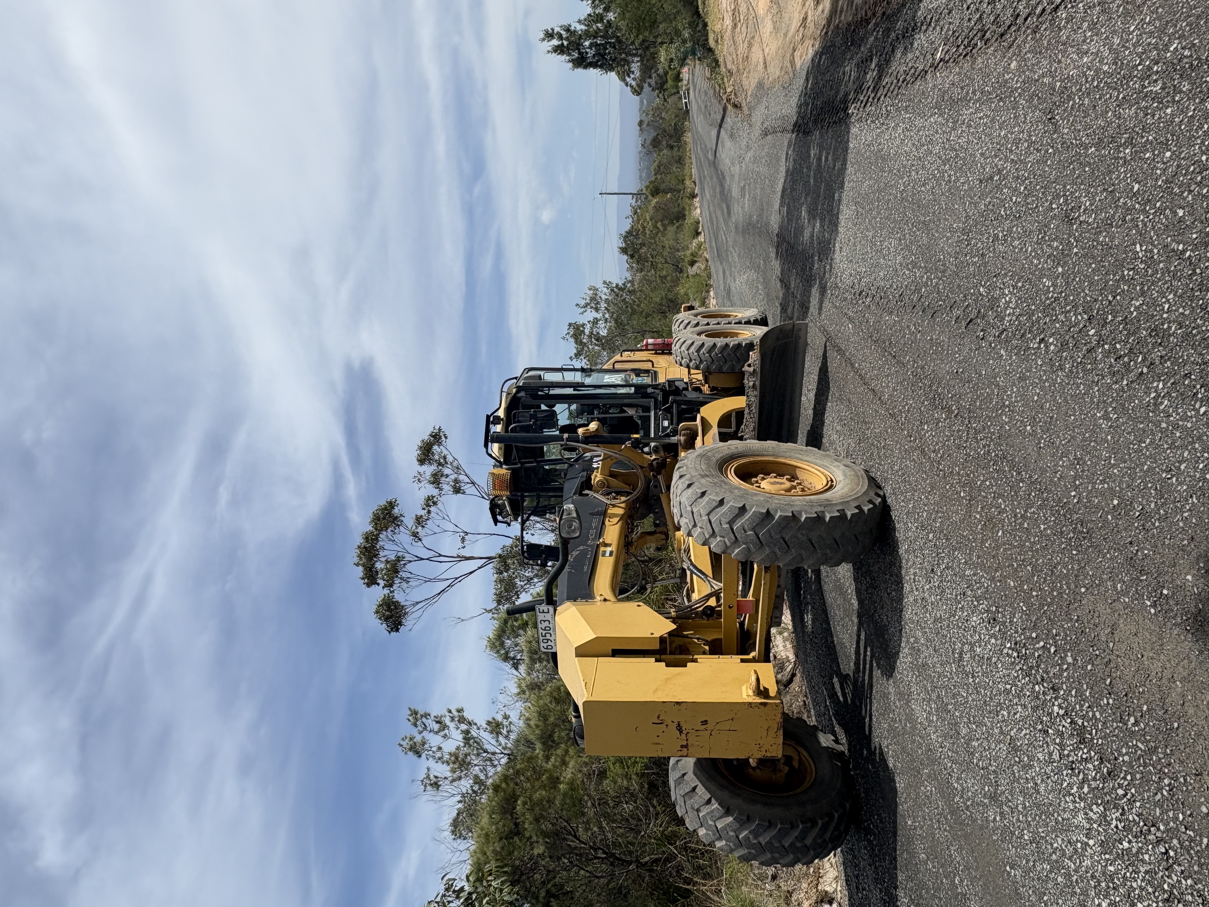 Yellow road grader parked on a gravel road under a partly cloudy sky with trees on the roadside.