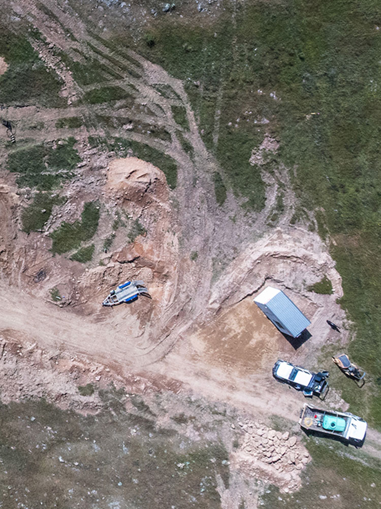 Aerial view of a dirt road with exposed earth, tire tracks, two parked vehicles, a small trailer, and various construction materials on grassy terrain.