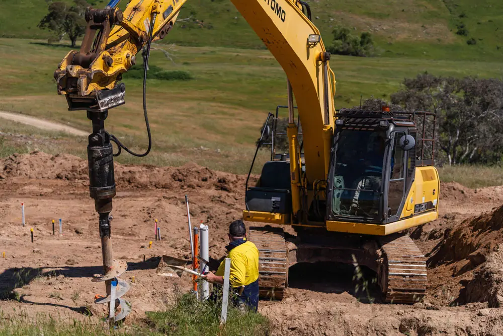 Construction worker in yellow shirt operating a yellow excavator with an auger drill attachment in a dirt field.