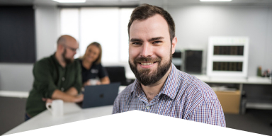 Smiling man with beard and plaid shirt in office with two colleagues working on a laptop in the background.