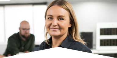 Smiling woman with blonde hair in an office setting with a man and electronic equipment blurred in the background.