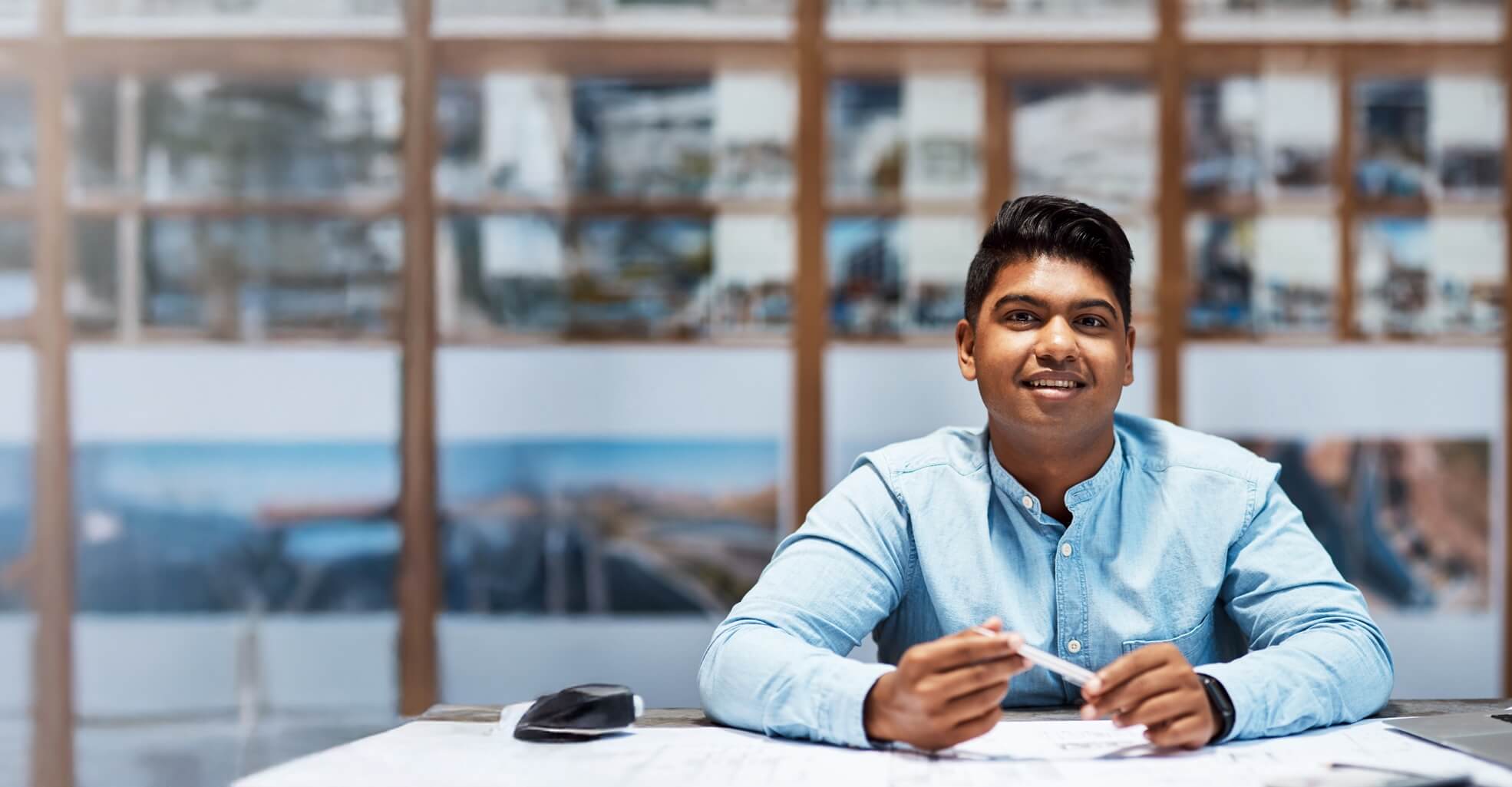 Smiling young man in a light blue shirt sitting at a table with architectural plans and holding a pen.