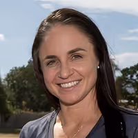 Smiling woman with long dark hair standing outdoors with trees and blue sky in the background.