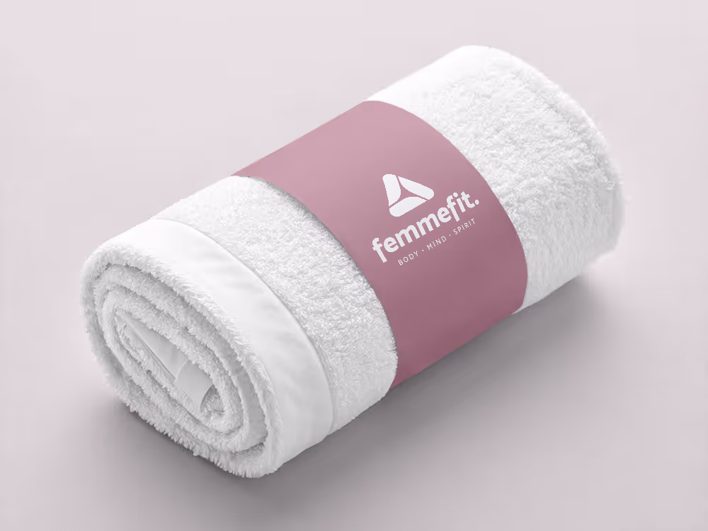 Rolled soft pink towel with a bright pink label that reads 'femmefit. BODY · MIND · SPIRIT' on a light background.