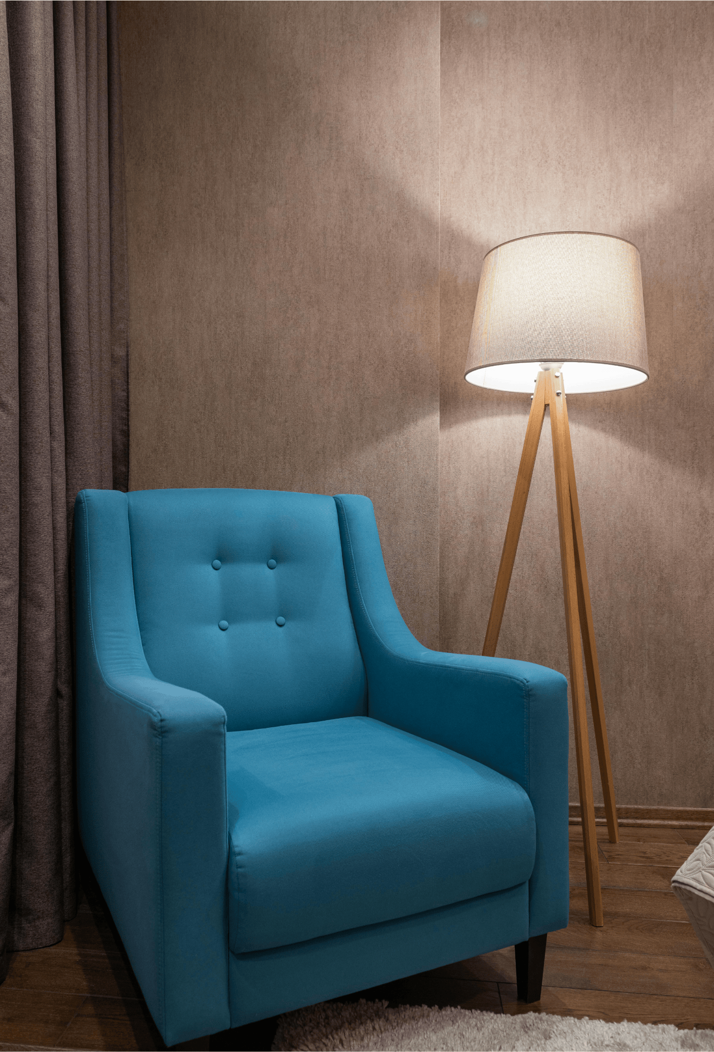 Blue upholstered armchair next to a wooden tripod floor lamp with a beige lampshade in a cozy room corner.