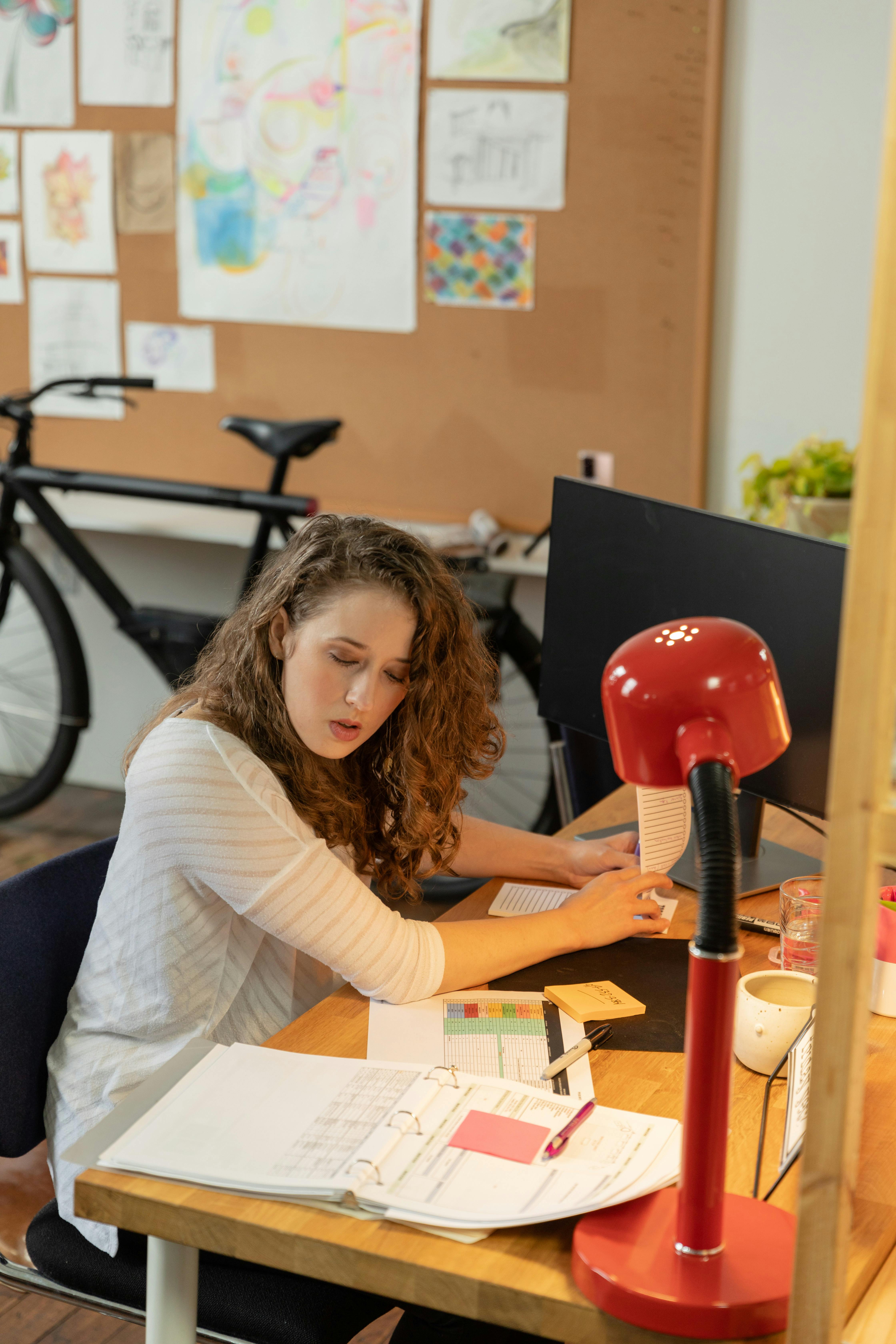 Woman sitting at a desk filled with documents, pens, and a red desk lamp, working on paperwork in a room with a bicycle and pinned drawings on the wall.