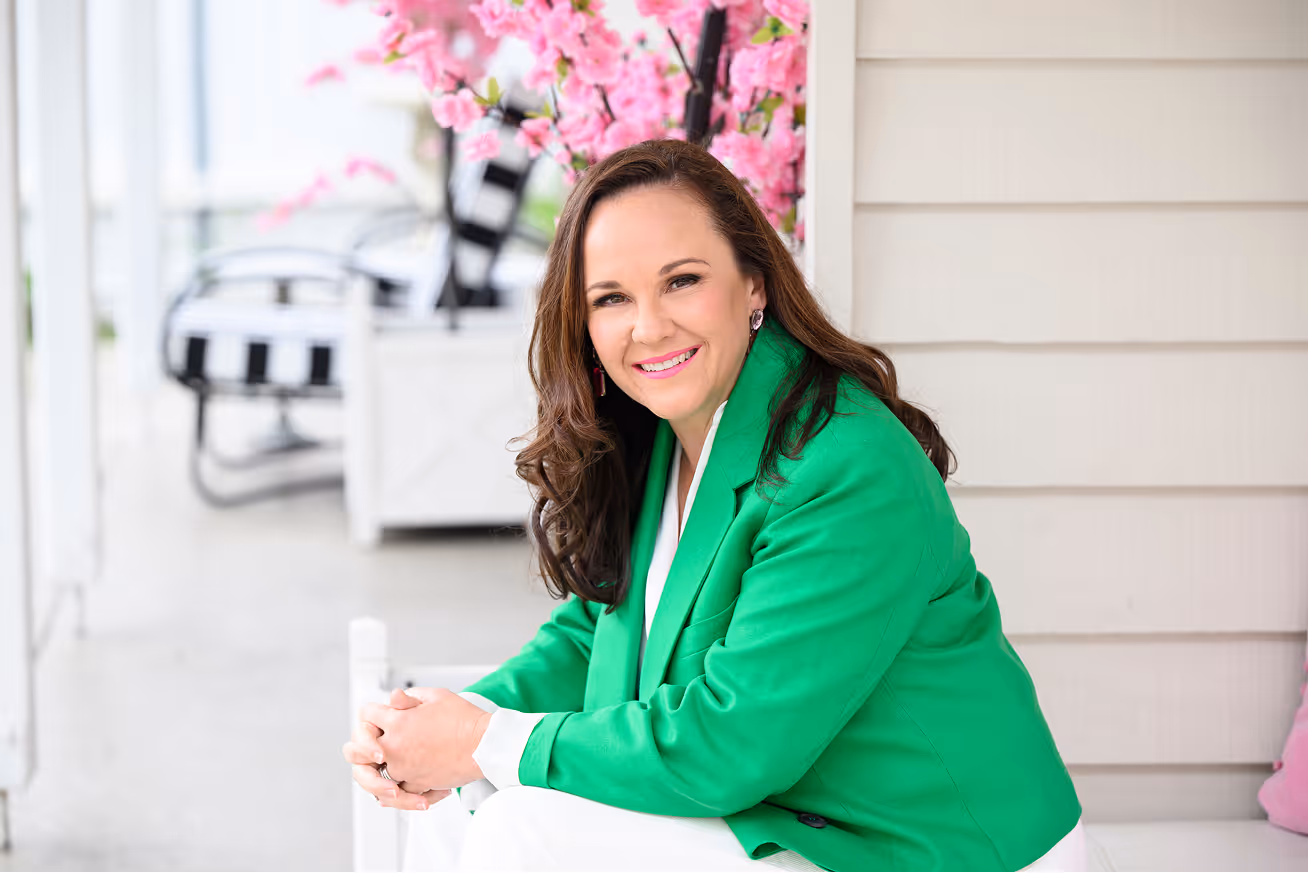 Smiling woman in green blazer sitting outdoors near pink flowering tree.