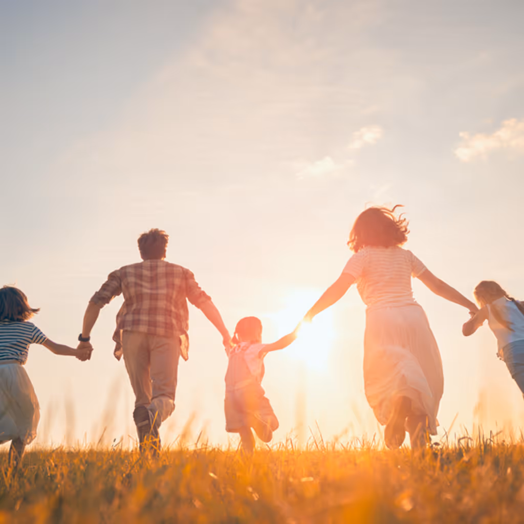 Family of two adults and three children running hand in hand through a sunlit grassy field at sunset.