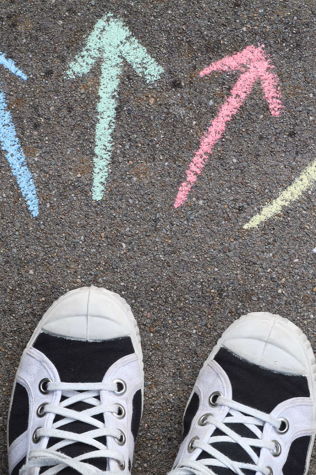Pair of white and black sneakers standing on asphalt with colorful chalk arrows pointing forward.