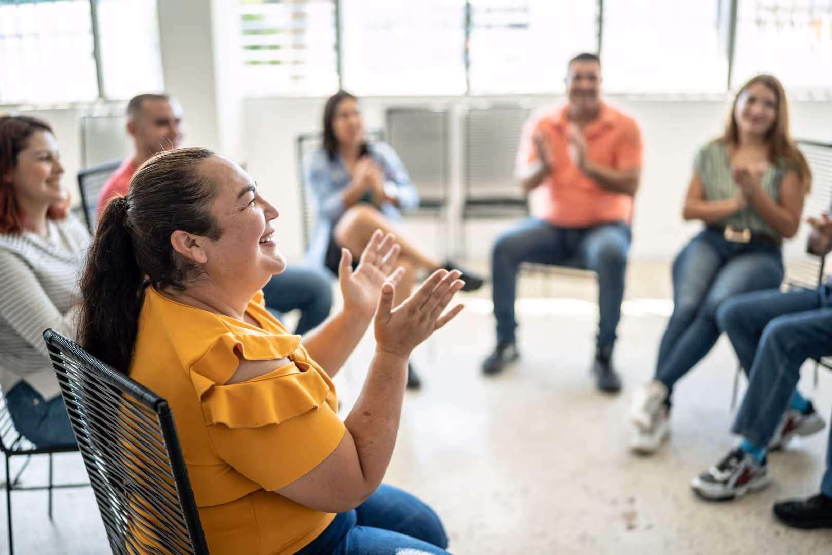 Smiling woman in yellow claps hands in a seated group therapy or workshop session.