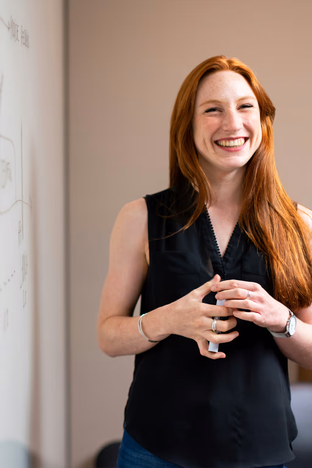 Smiling red-haired woman holding a marker stands near a whiteboard with writing.