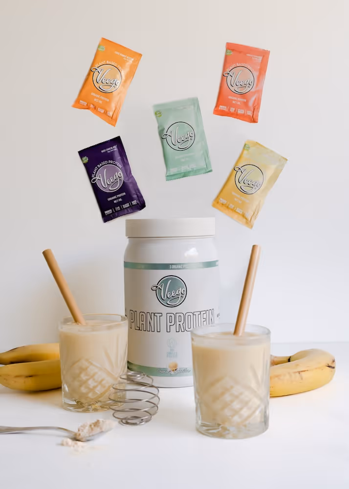 Veego plant protein powder container with five colorful sample packets and two banana smoothies in glasses.