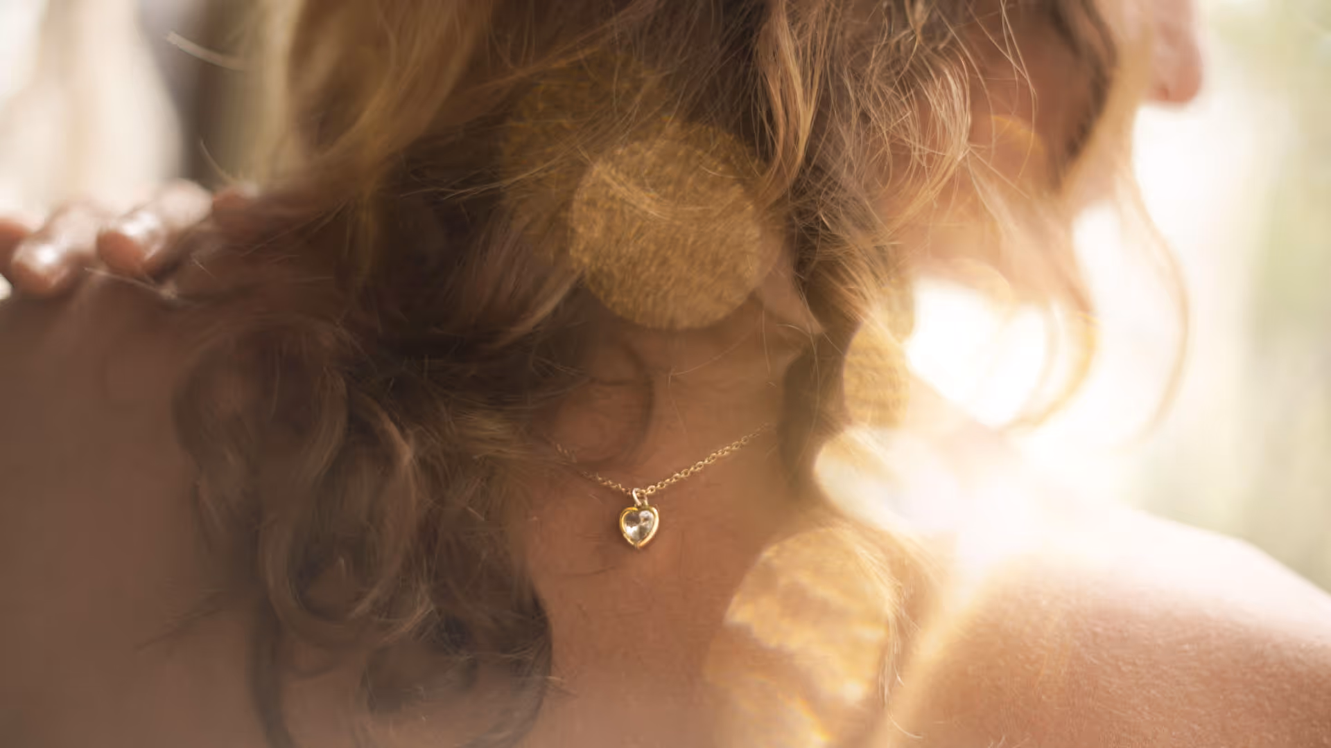 Close-up of a woman wearing a gold heart-shaped pendant necklace with soft sunlight effects.