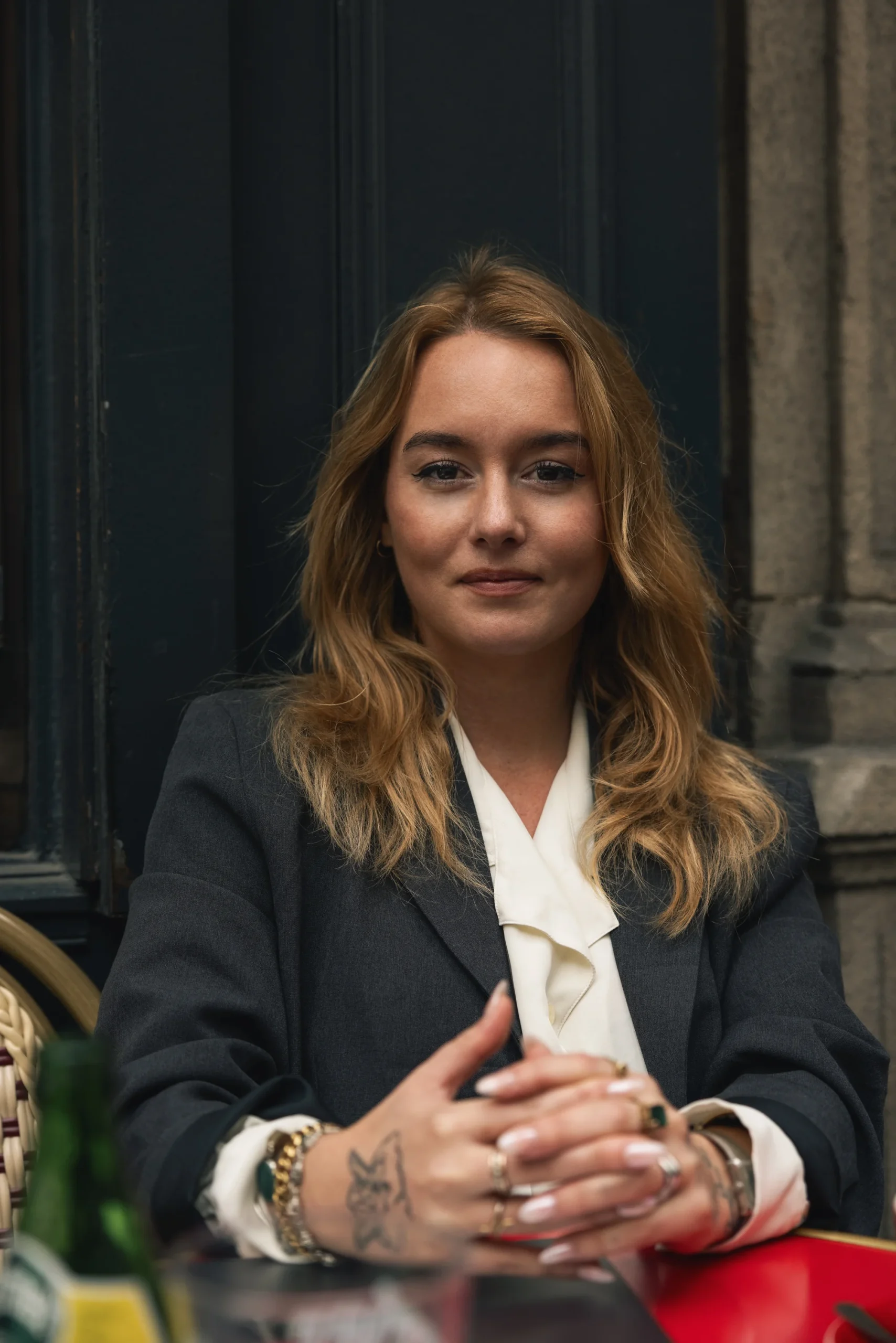 Confident woman with blonde hair wearing a dark blazer and white blouse sitting at a table with hands clasped.