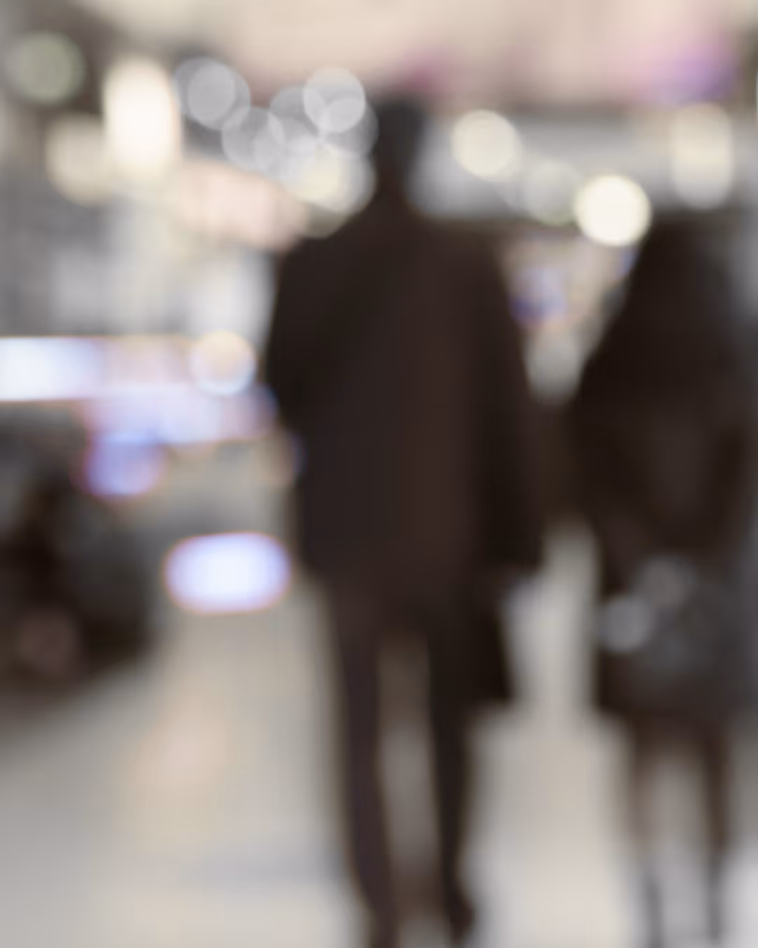 Blurred image of two people walking indoors with bright lights in the background.