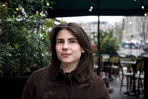 Black and white photo of a woman in a white blazer looking back with a blurred background of a grid window and textured wall.