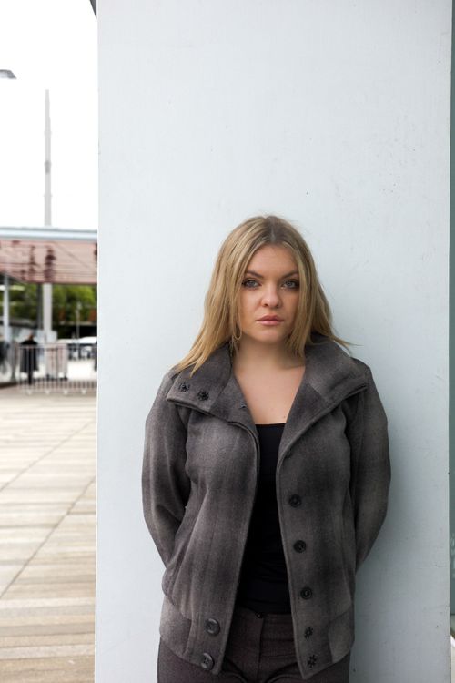 Black and white photo of a woman in a white blazer looking back with a blurred background of a grid window and textured wall.