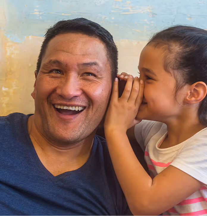 Smiling man listening as a young girl whispers into his ear indoors.