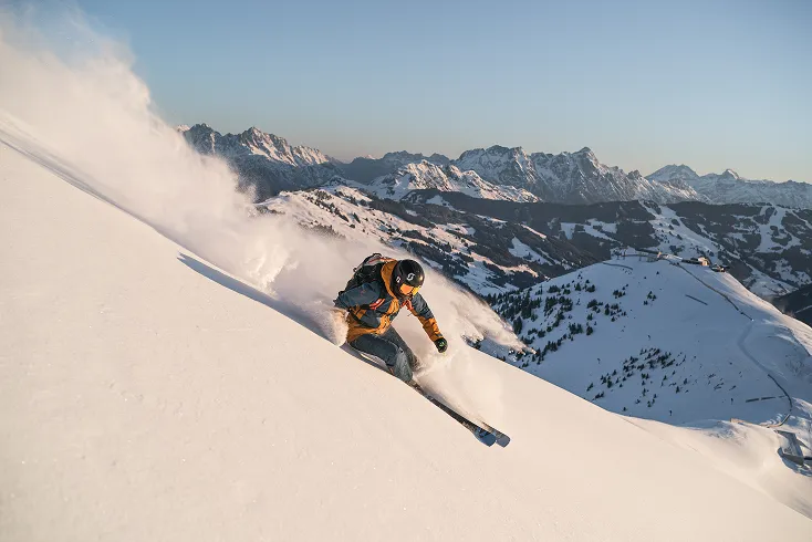 Skier making a sharp turn on a snow-covered mountain slope with snowy peaks in the background.