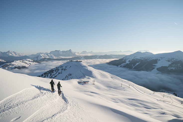 Two skiers walk on a snowy ridge with ski lifts and mountains visible above a sea of clouds under a clear blue sky.