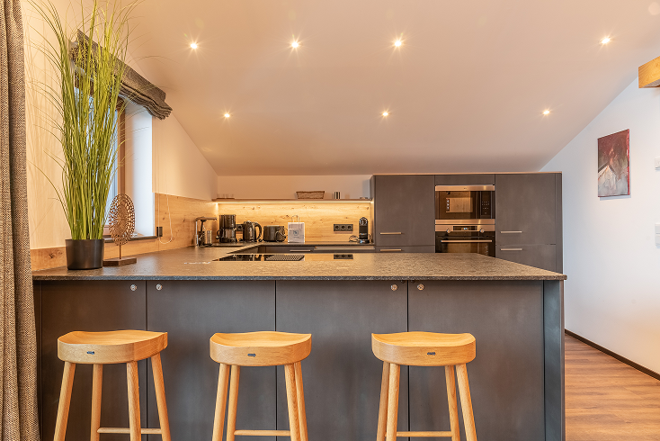 Modern kitchen interior with a large dark island countertop, three wooden stools, built-in ovens, and a tall potted plant by the window.