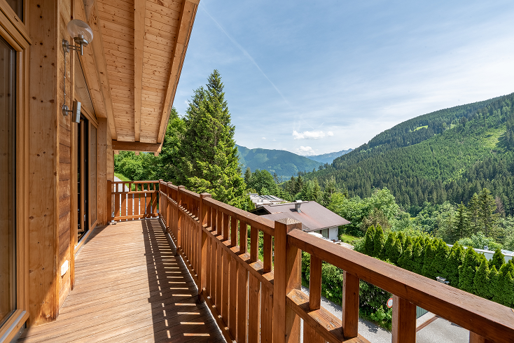 Wooden balcony of a mountain cabin overlooking green forested hills under a partly cloudy blue sky.