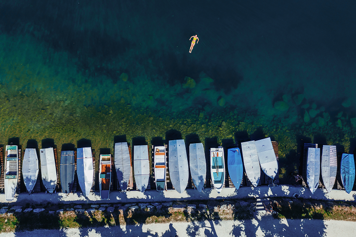 Aerial view of a person floating on their back in clear blue water near a row of docked boats along a shoreline.