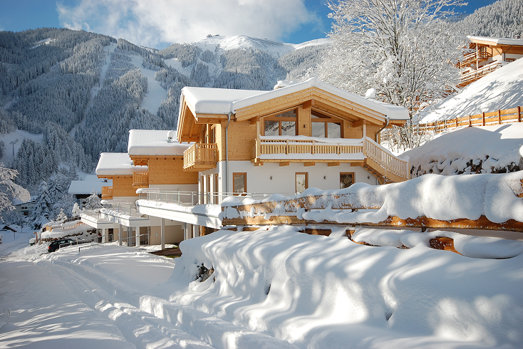Wooden chalet houses covered in snow with a snowy mountain and trees in the background under a blue sky.