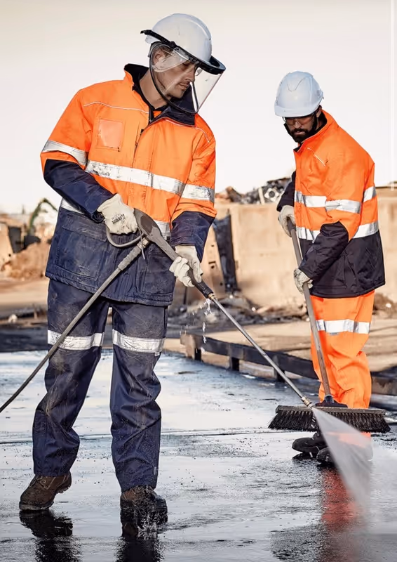 Two construction workers in orange and navy high-visibility gear cleaning a wet surface using a pressure washer and a broom.