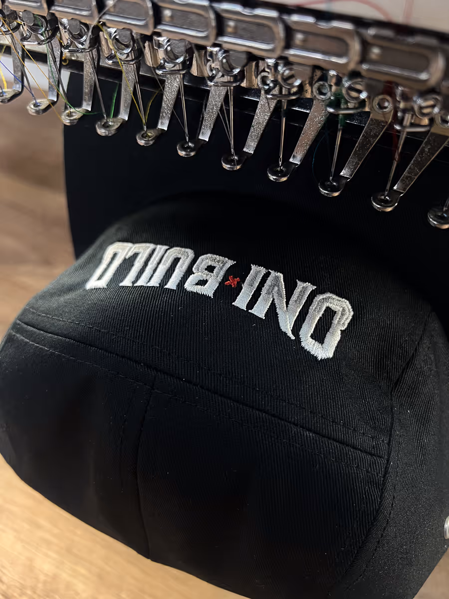 Embroidery machine stitching white and red text on a black cap reading 'ONI BULLI'.