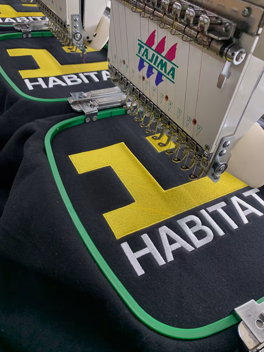 Industrial embroidery machine stitching a yellow and white 'HABITAT' logo on black fabric framed by green embroidery hoops.
