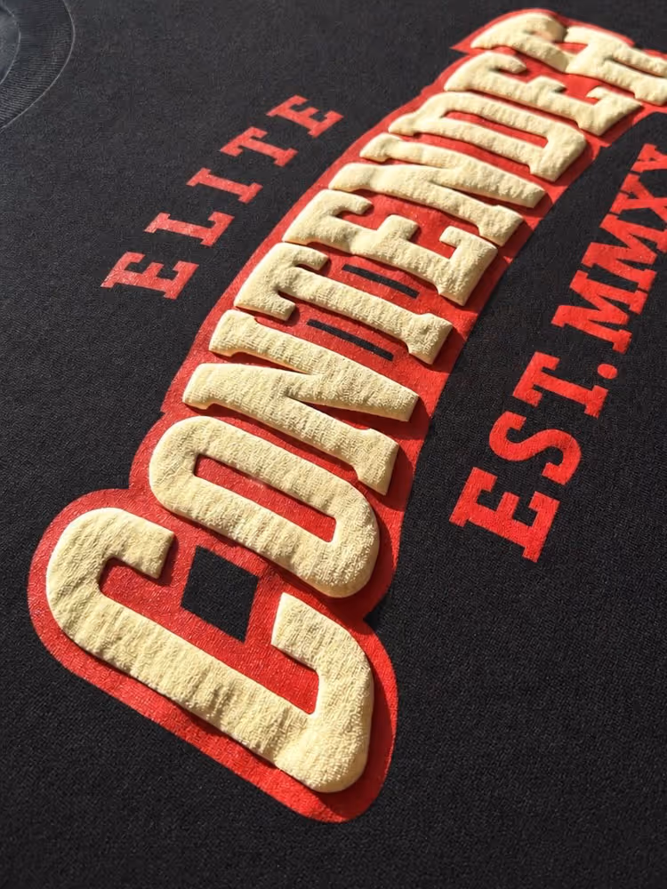 Close-up of black shirt with raised beige text reading COVDENDEE, red text ELITE and EST. MMXX.