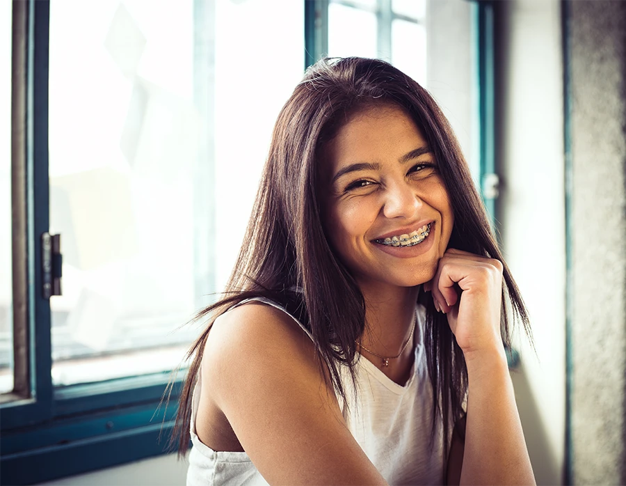 Smiling young woman with long dark hair and braces sitting indoors by a window.