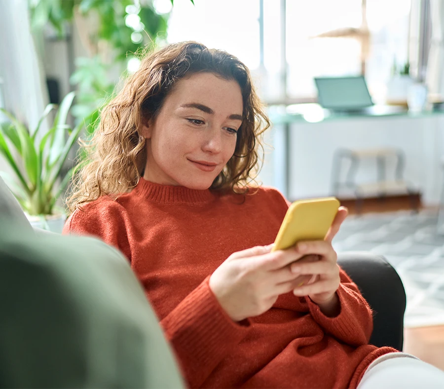 Woman with curly hair sitting on a couch at home, smiling while looking at a yellow smartphone.