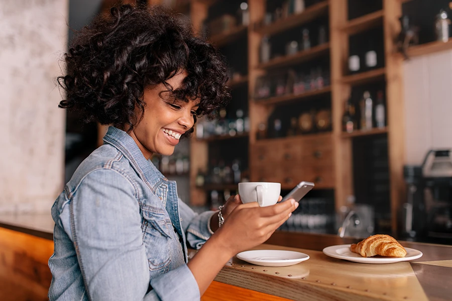 Smiling woman in a denim jacket sitting at a cafe counter holding a smartphone and a white coffee cup, with a croissant on a plate nearby.