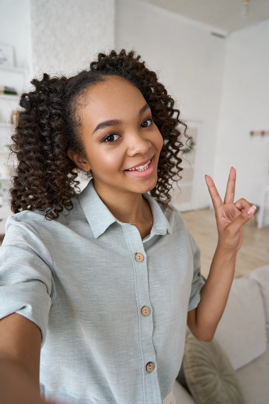 Smiling teenage girl with curly hair taking a selfie and making a peace sign with her hand.