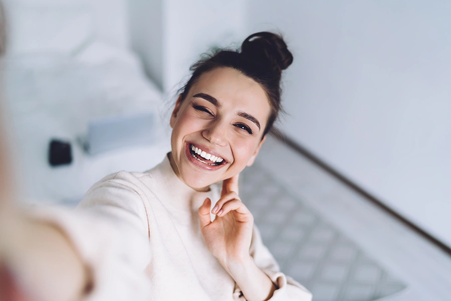 Smiling young woman with a top bun hairstyle taking a playful selfie while winking and touching her cheek.
