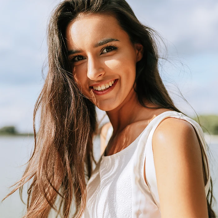 Young woman with long brown hair smiling outdoors by a body of water under a partly cloudy sky.