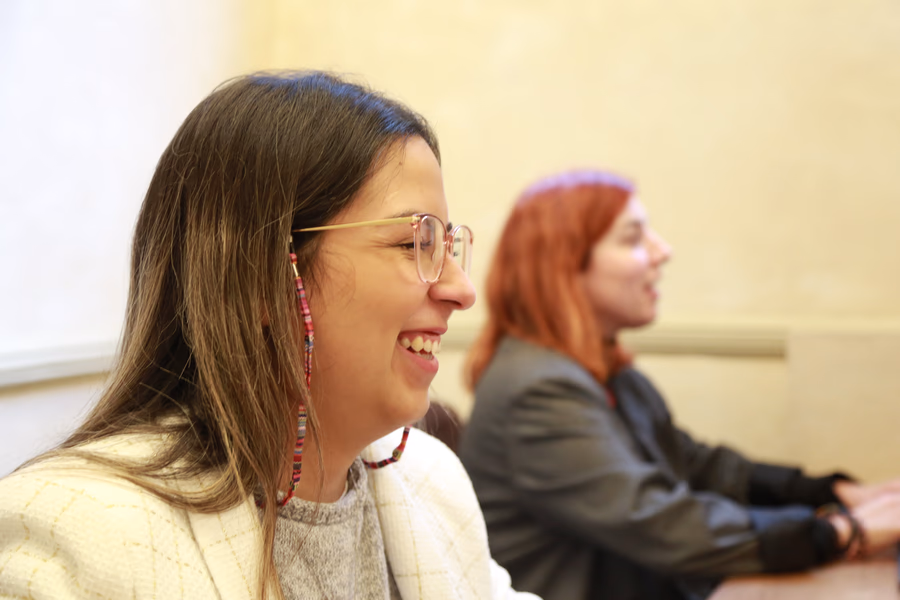 Two women sitting side by side and smiling, one wearing glasses and a white jacket, the other with red hair and a dark jacket.